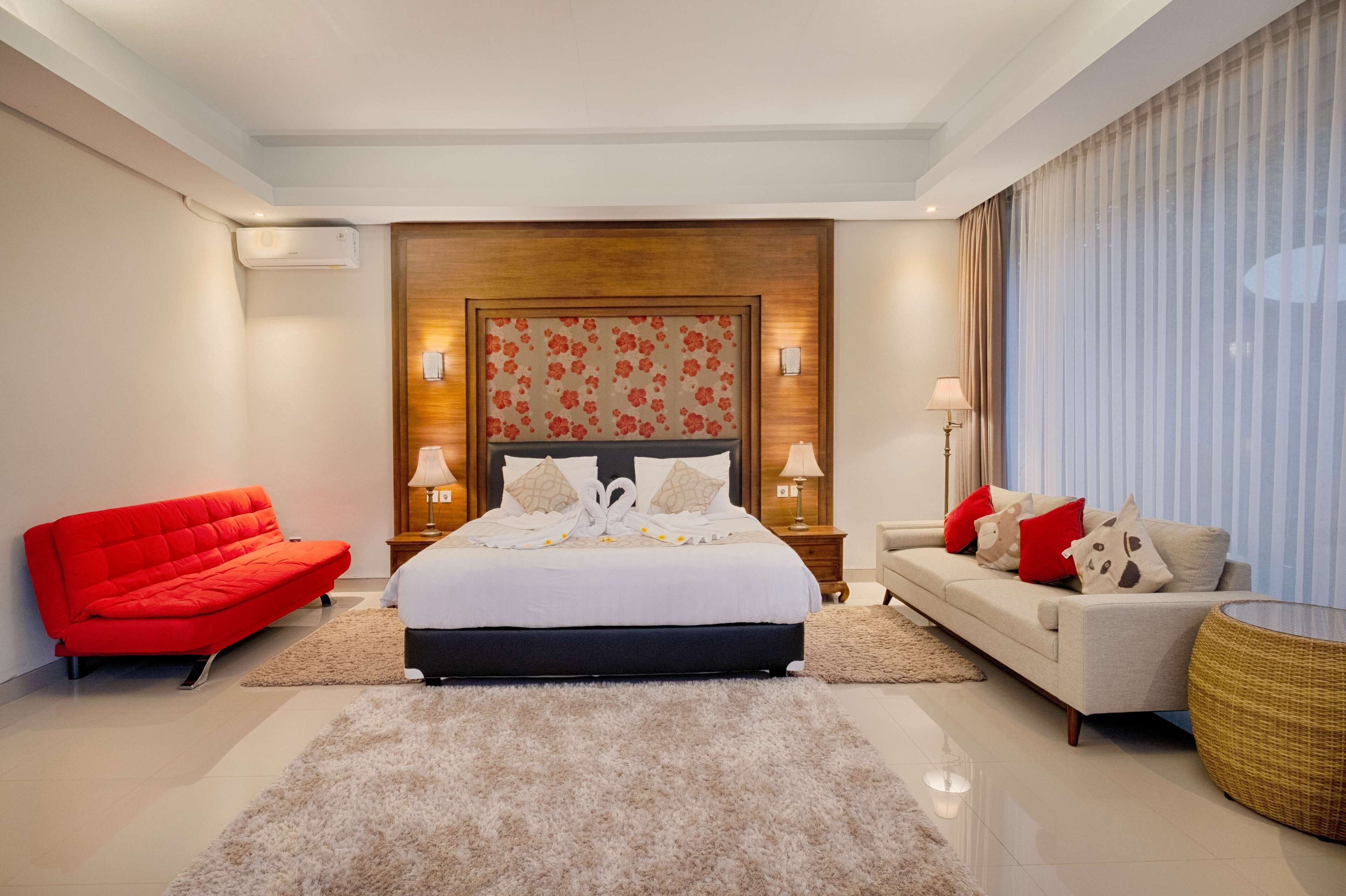 A modern hotel room with a large bed, a red sofa, a white sofa, and a round wicker table.