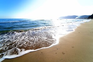 A beach with waves gently lapping at the shore, clear skies, and distant mountains. - B&B My Home (Alassio)