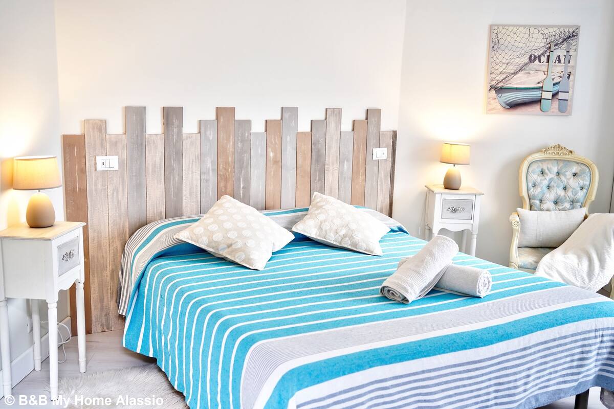 A neatly arranged bedroom with a wooden headboard, striped bedding, a decorative wall piece, and a classic chair.
