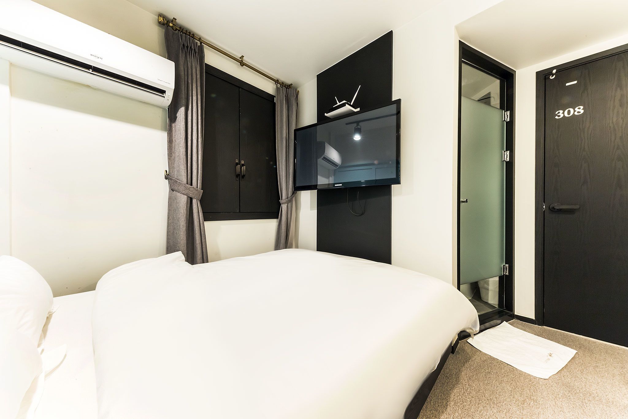 A hotel room with a bed, a flat-screen TV mounted on the wall, a door with the number 308, and a window with curtains.