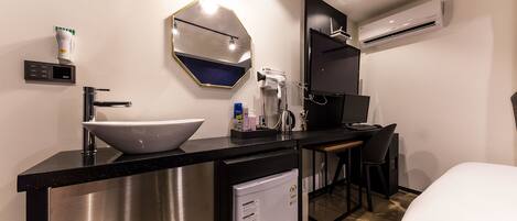 A compact hotel room with a modern kitchenette, a white sink, a compact refrigerator, a small dining table, and a single bed.