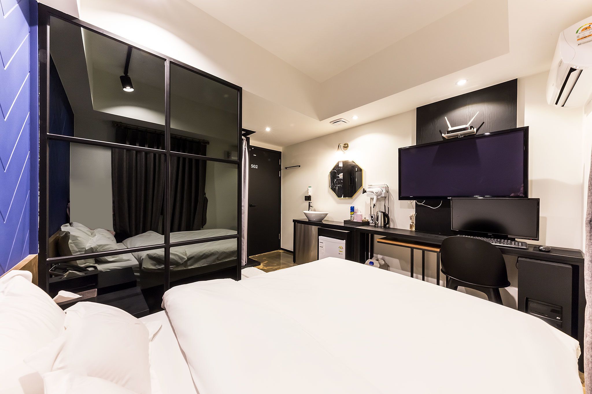A modern hotel room with a large bed, a desk with a computer, a TV, and a bathroom with a shower.