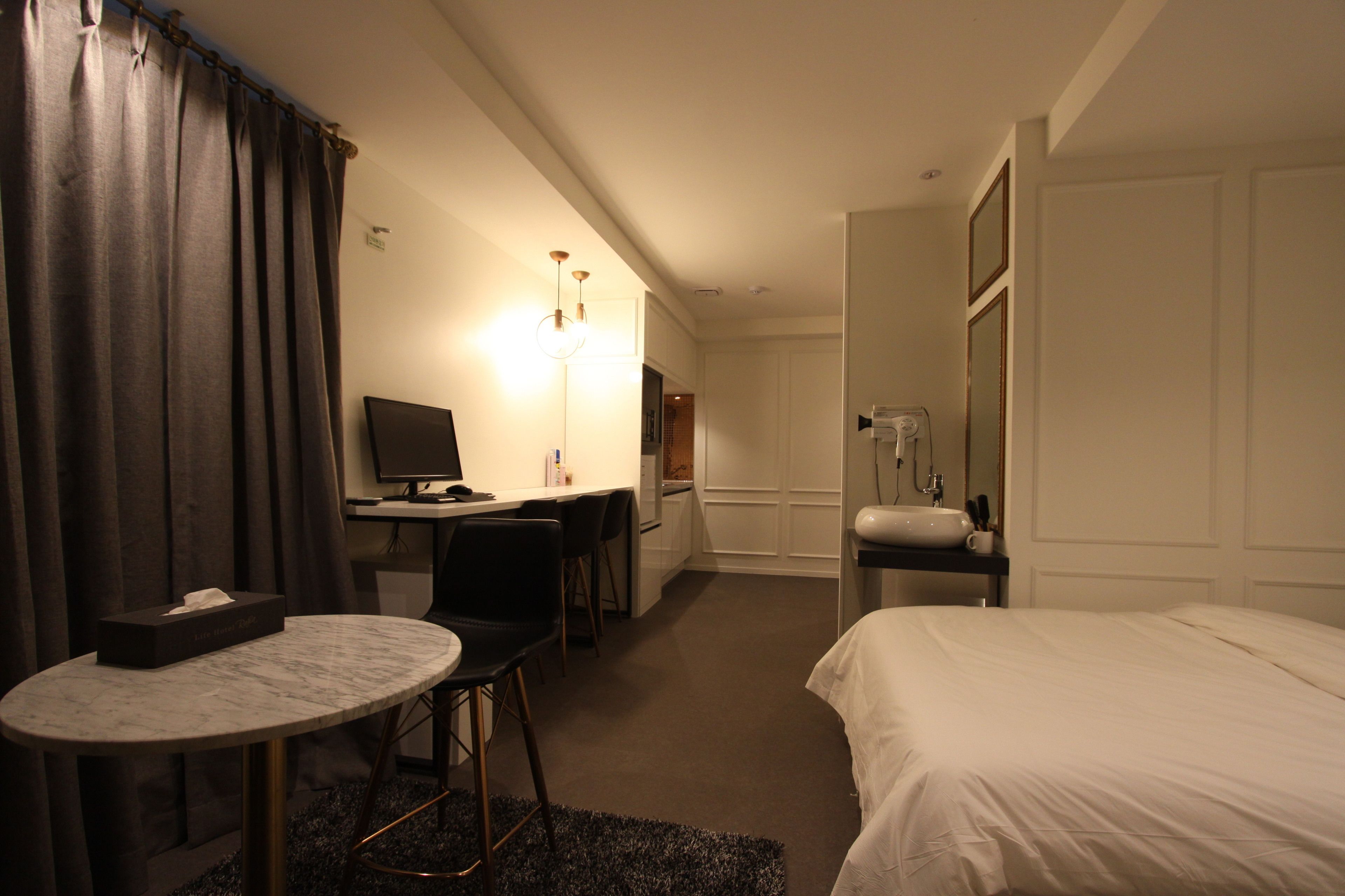 Une chambre d’hôtel comprenant un lit, un bureau avec un ordinateur, une chaise, une petite table, une lampe et une salle de bain avec un lavabo et un miroir.