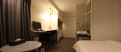 Une chambre d’hôtel comprenant un lit, un bureau avec un ordinateur, une chaise, une petite table, une lampe et une salle de bain avec un lavabo et un miroir.