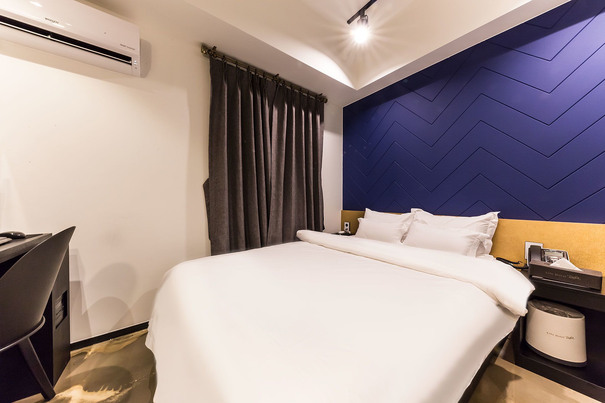 A hotel room with a bed, a desk, an air conditioner, a nightstand, and a lamp.