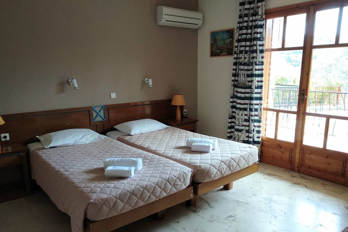 Ein Doppelzimmer mit zwei Betten, jeweils mit einer gesteppten Bettdecke und zwei Handtüchern. Es gibt einen Nachttisch mit einer Lampe, eine Klimaanlage an der Wand und ein Gemälde über dem Bett.