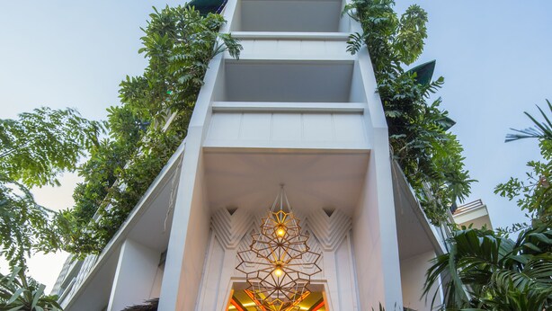 A modern hotel entrance with a geometric facade, indoor seating area, and lush greenery. - Natural House (Siem Reap)