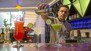 A bartender pouring a drink into a glass with a lime wedge. There are three other drinks on the bar counter. - Natural House (Siem Reap)