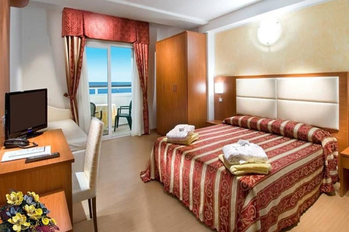 A hotel room with a bed, a desk with a computer, a chair, a TV, and a view of the sea.