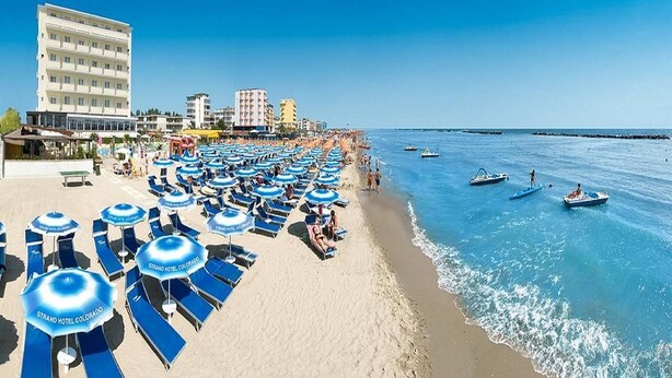 Una playa con filas de tumbonas y sombrillas, un edificio de hotel y barcos en el agua.