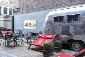 Une caravane ancienne est garée devant un bâtiment en briques, sur lequel on peut voir une enseigne indiquant la présence d’un bar à vin et d’un hôtel. Des espaces de détente extérieurs, avec des chaises et des tables, sont aménagés.