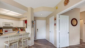 A kitchen with white cabinets, a microwave, and a stove. There is a bar area with a chair and a small table. The floor is wooden, and there are two white doors in the hallway.