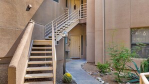 A building with a staircase leading to an upper floor, a door, and a small garden with a cactus and rocks.