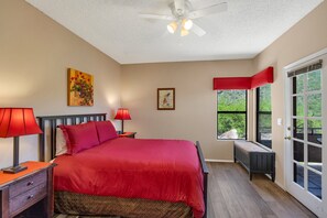 A bedroom with a bed, two nightstands, a ceiling fan, a window with a view, and a door.