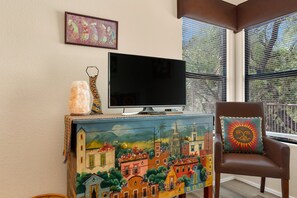 Ein Wohnzimmer mit Fernseher auf einer Kommode, einem Sessel mit Sonnenkissen und einem farbenfrohen Wandgemälde.