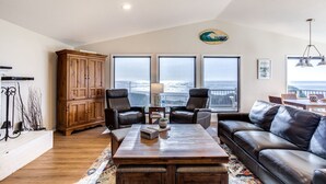 A spacious living room with a wooden cabinet, leather chairs, a wooden coffee table, and a leather sofa. Large windows offer a view of the ocean.
