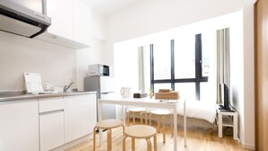 A modern kitchen with white cabinets, a microwave, and a coffee maker. There are three stools at a table, and a bed with a TV in the background. - OH Inn -Fukuoka Stay- (Fukuoka)