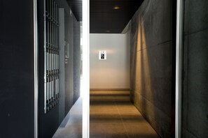 Un couloir étroit au design moderne, avec une cloison en verre d’un côté et un mur en béton de l’autre. Un petit panneau de commande rétroéclairé est visible sur le mur.