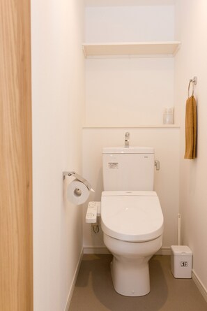 Une petite salle de bain propre, avec des toilettes blanches, un porte-rouleau de papier toilette et une petite poubelle.