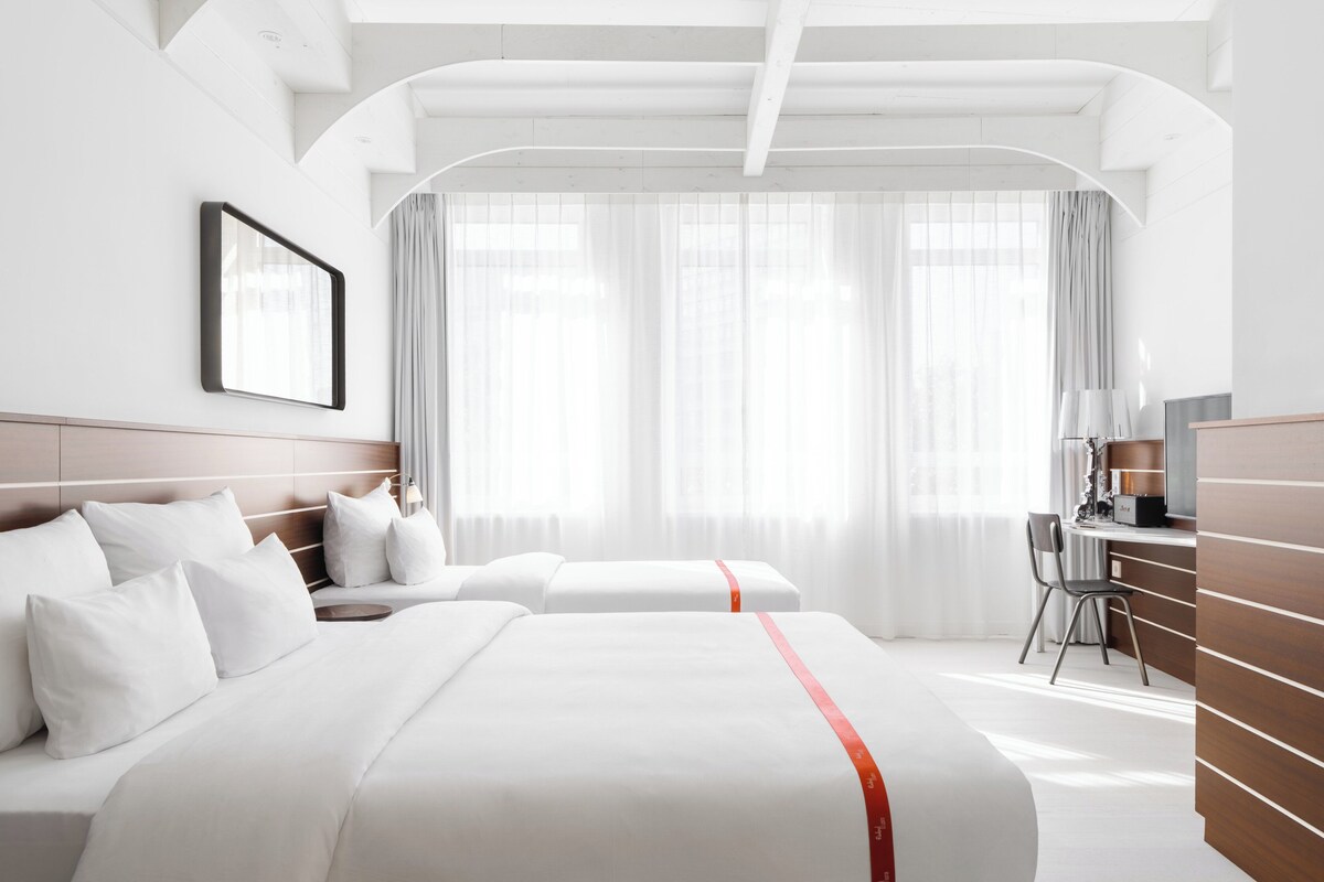A hotel room with two beds, a desk, a chair, and a large window with curtains.