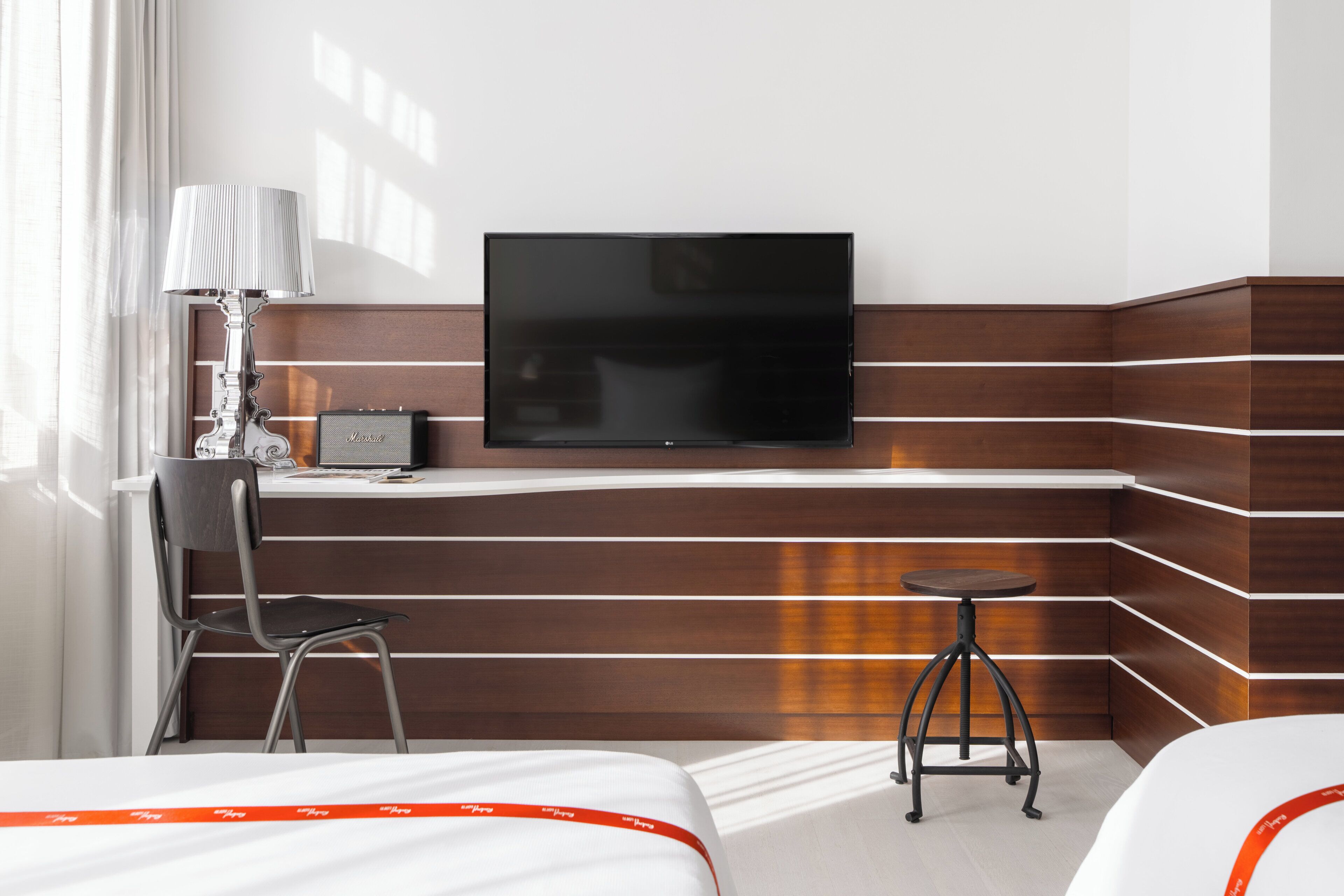 Une chambre d’hôtel comprenant un lit, un bureau avec une chaise, un téléviseur fixé au mur et un petit tabouret.
