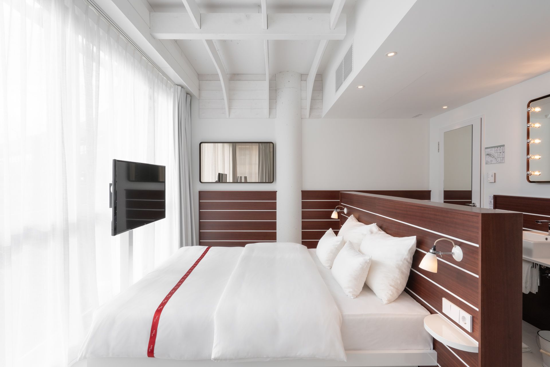 A modern hotel room with a large bed, a television, a mirror with lights, and a bathroom with a sink and mirror.
