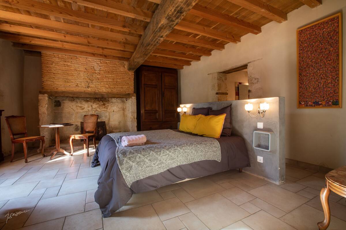 A bedroom with a bed, wooden ceiling beams, a brick wall, a small table with chairs, and a painting on the wall.