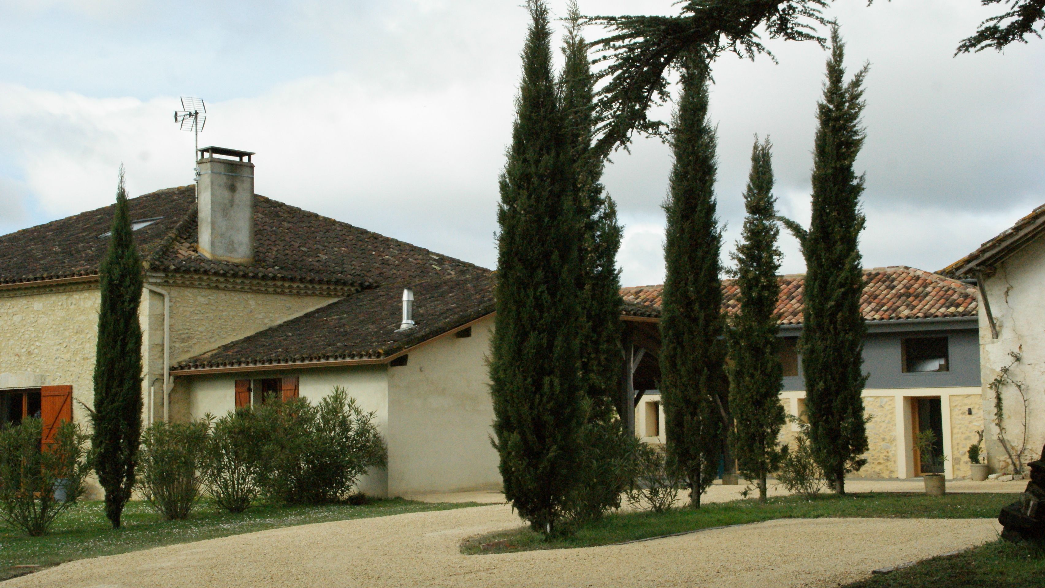 A traditional house with a tiled roof, a chimney, and a gravel driveway. There are tall evergreen trees in the front yard.