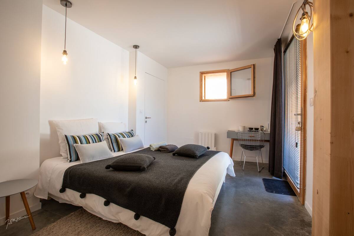 A bedroom with a bed, bedside table, and a chair. There is a window with blinds and a radiator on the wall.