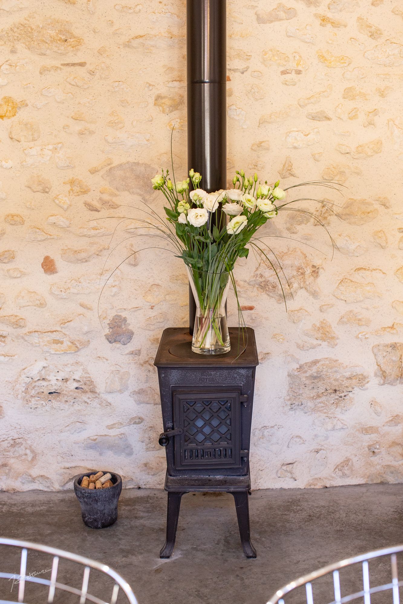 A rustic stove with a vase of white flowers on top, a bucket of wine corks, and a metal chair in front.