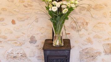 A rustic stove with a vase of white flowers on top, a bucket of wine corks, and a metal chair in front.