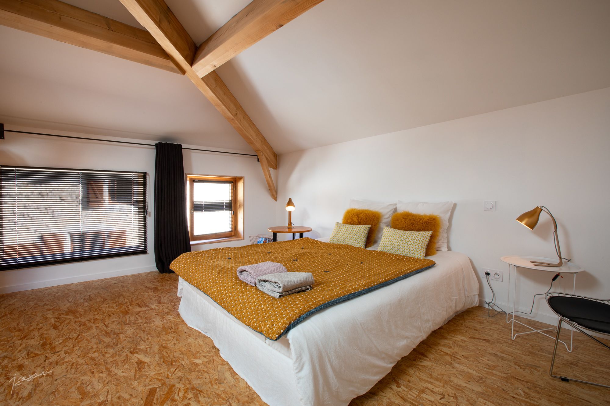 Une chambre avec un plafond en pente, un lit recouvert d’une couverture à motifs, deux oreillers, une table de chevet avec une lampe et une fenêtre avec des stores.