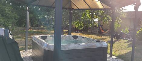 A hot tub under a covered patio with a pergola, surrounded by gravel and greenery.
