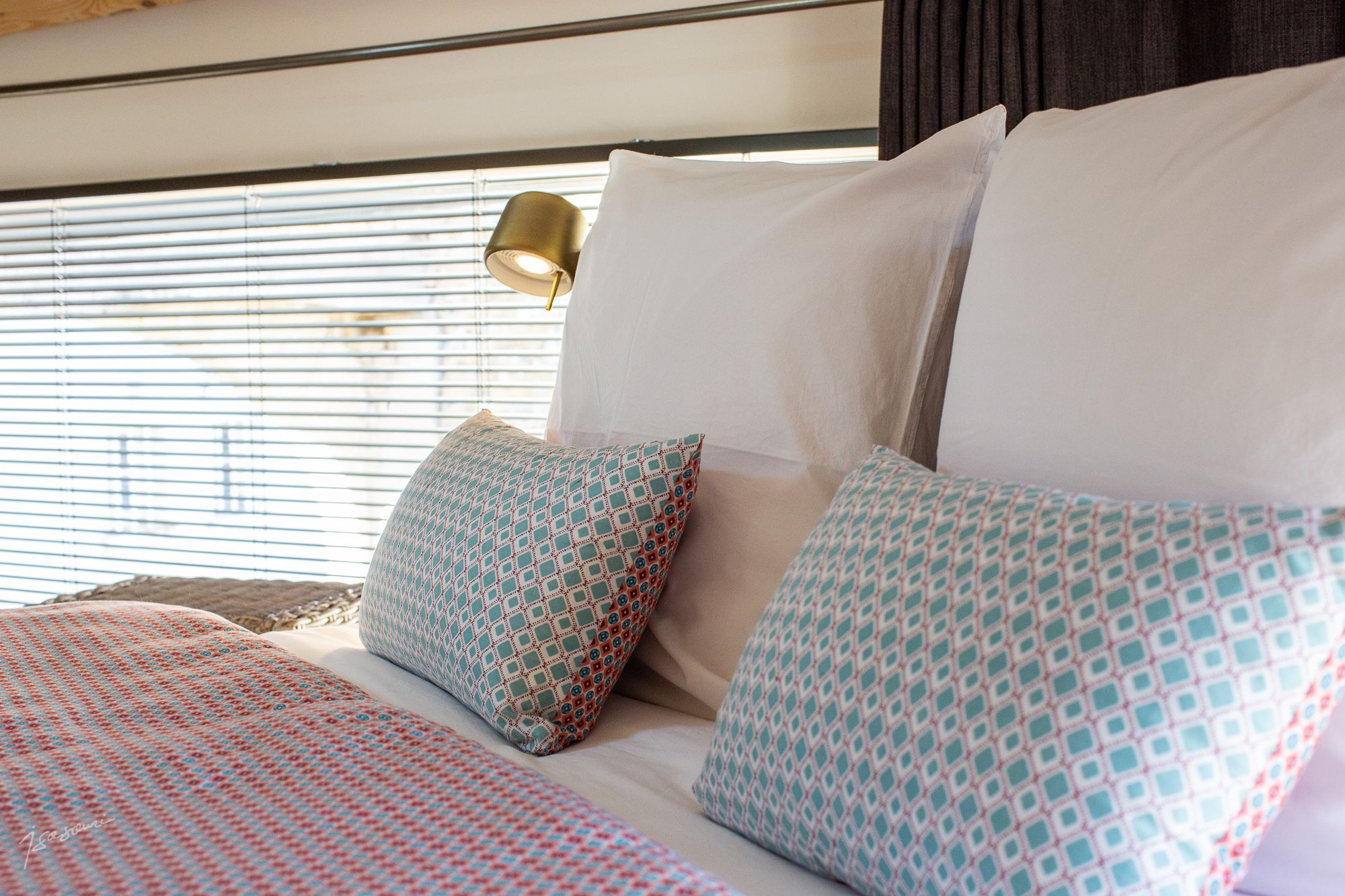 A neatly made bed with patterned pillows and a plain white pillow, a gold lamp on a bedside table, and a window with blinds.