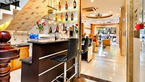 A bar area with a counter, bar stools, and shelves with bottles. There is a staircase with a patterned wall and a chandelier. - HANZ MinhVy Hotel (Ho Chi Minh City)