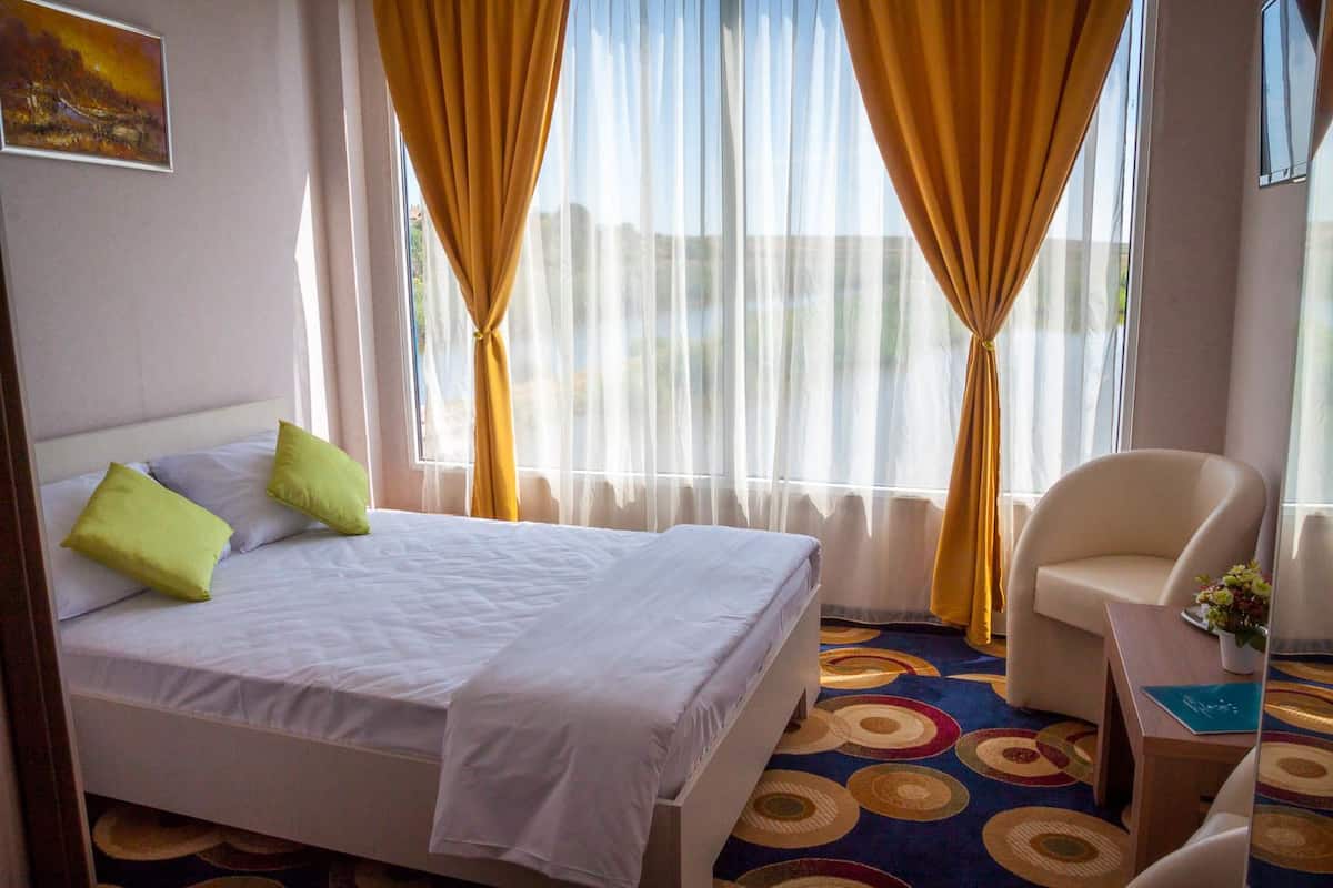 A hotel room with a bed, two pillows, a chair, a small table with a vase, and a window with curtains.