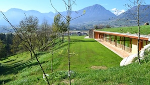 A modern building with a long glass facade, set against a backdrop of mountains and a grassy landscape. - Casariga (Comano Terme)