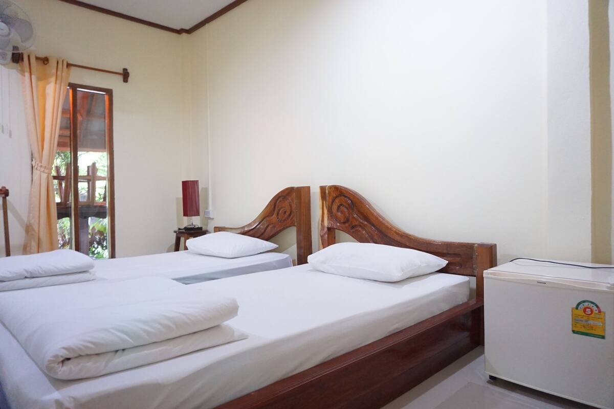 A room with two wooden beds, white bedding, a small table with a lamp, and a white refrigerator.