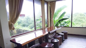 Una habitación con una larga mesa de madera y taburetes a juego, ventanas grandes con cortinas y vistas a un exuberante follaje.