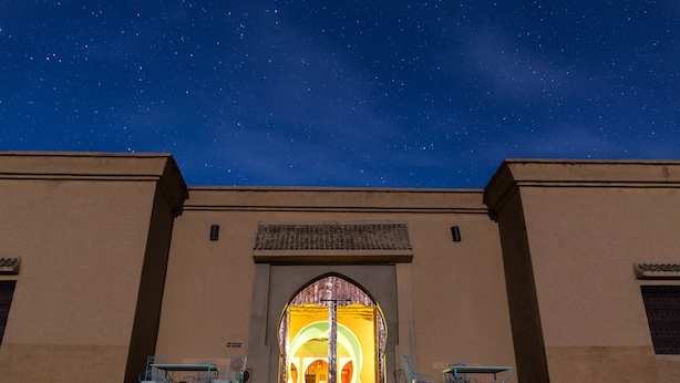 A building with a large arched entrance, two blue chairs outside, and a starry night sky. - Dar Mari (Taouz)