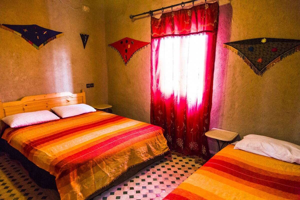 A room with a large bed, a smaller bed, a window with red curtains, and a checkered floor.