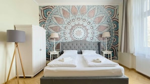 Una cama doble con cabecera tapizada de gris, dos mesitas de noche con lámparas, y un tapiz decorativo en la pared.