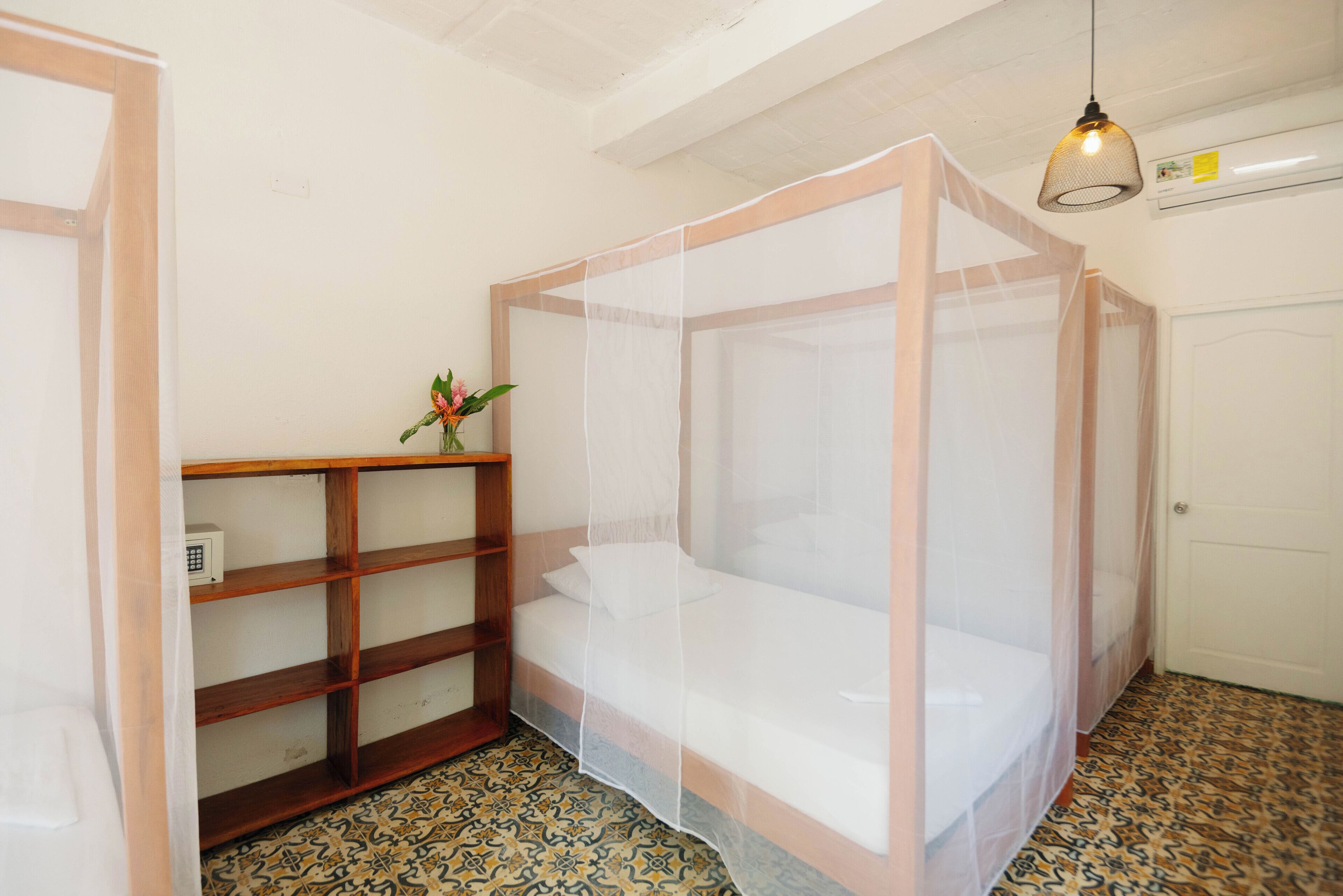 A room with a bed under a mosquito net, a wooden shelf with a vase of flowers, and patterned floor tiles.