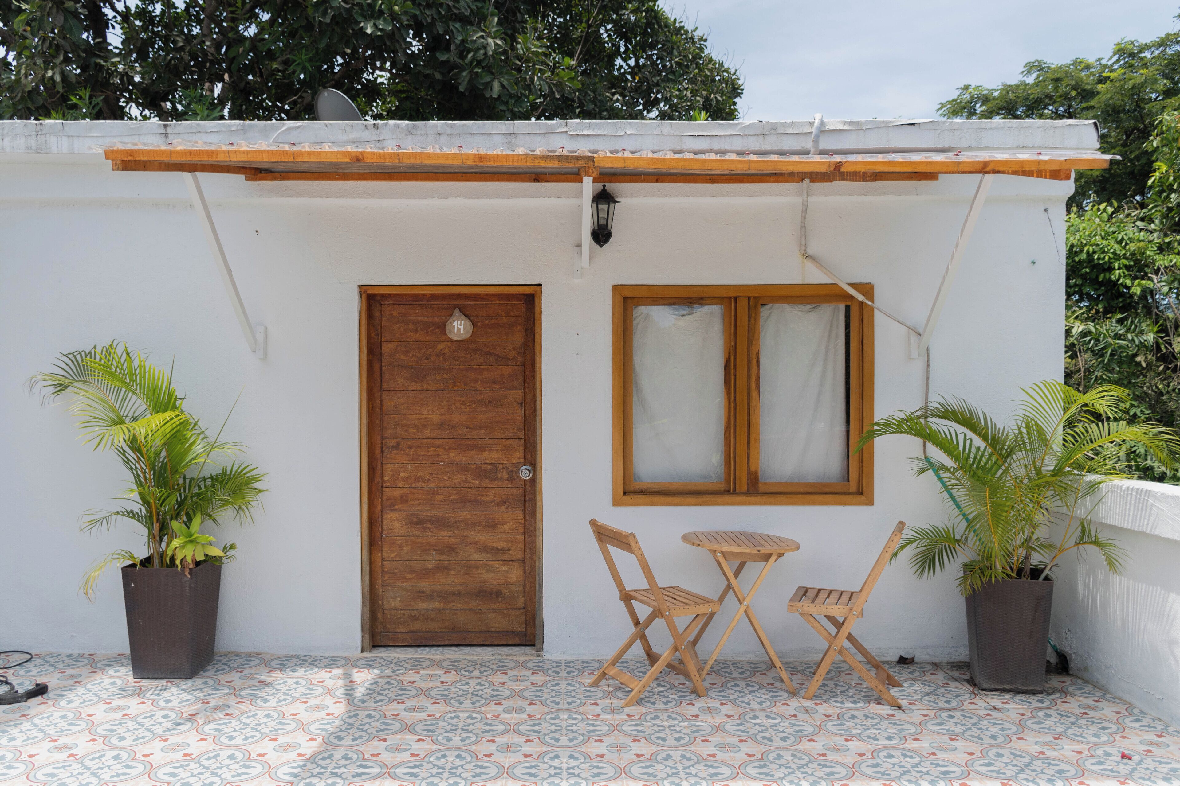 A small, white building with a wooden door and window, two wooden chairs, and two potted plants.