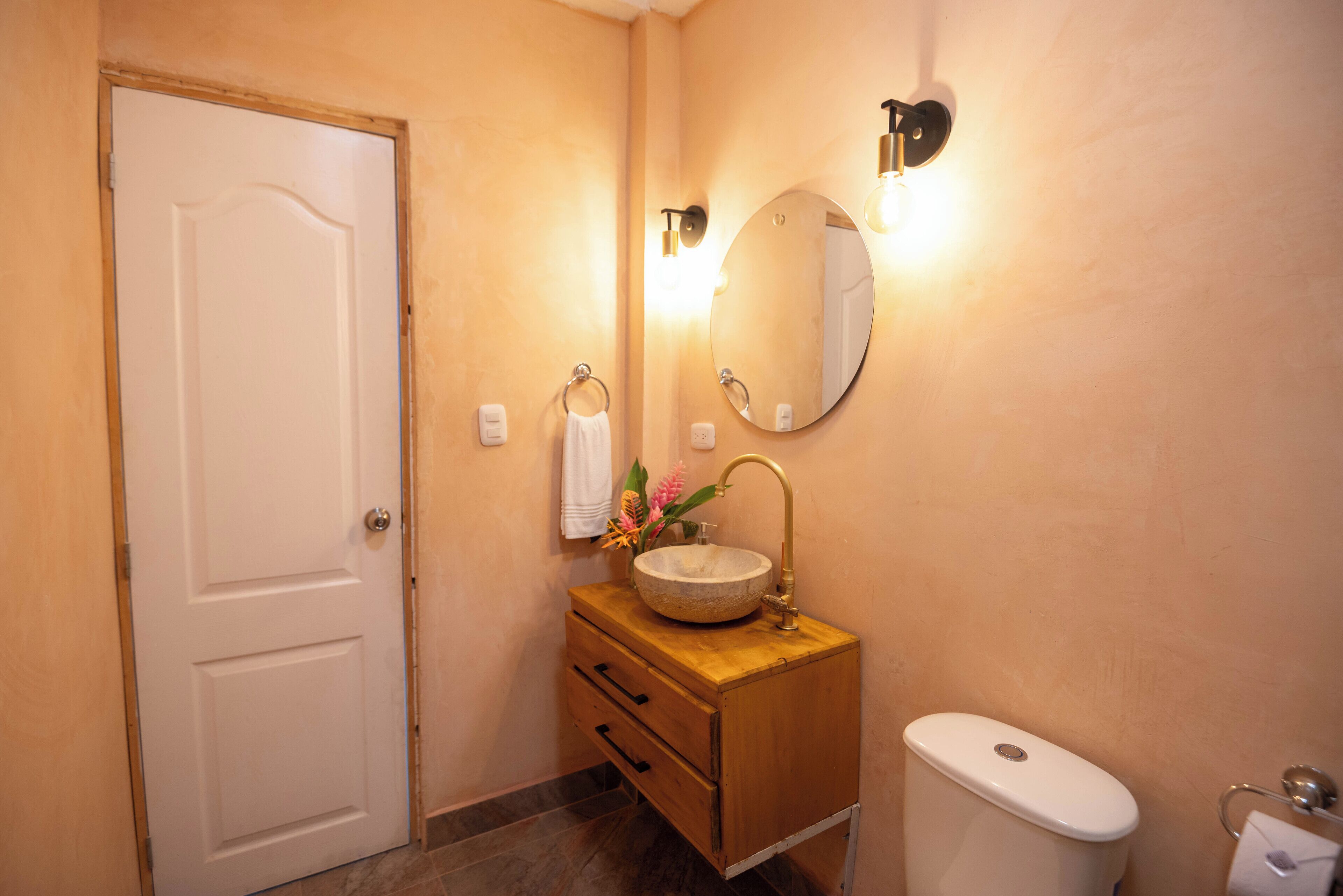 A bathroom with a wooden vanity, a round sink, a toilet, and a mirror with wall-mounted lights.