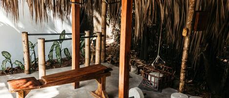 A room with a thatched roof, a wooden bench, a barbell, and weight plates on the floor.