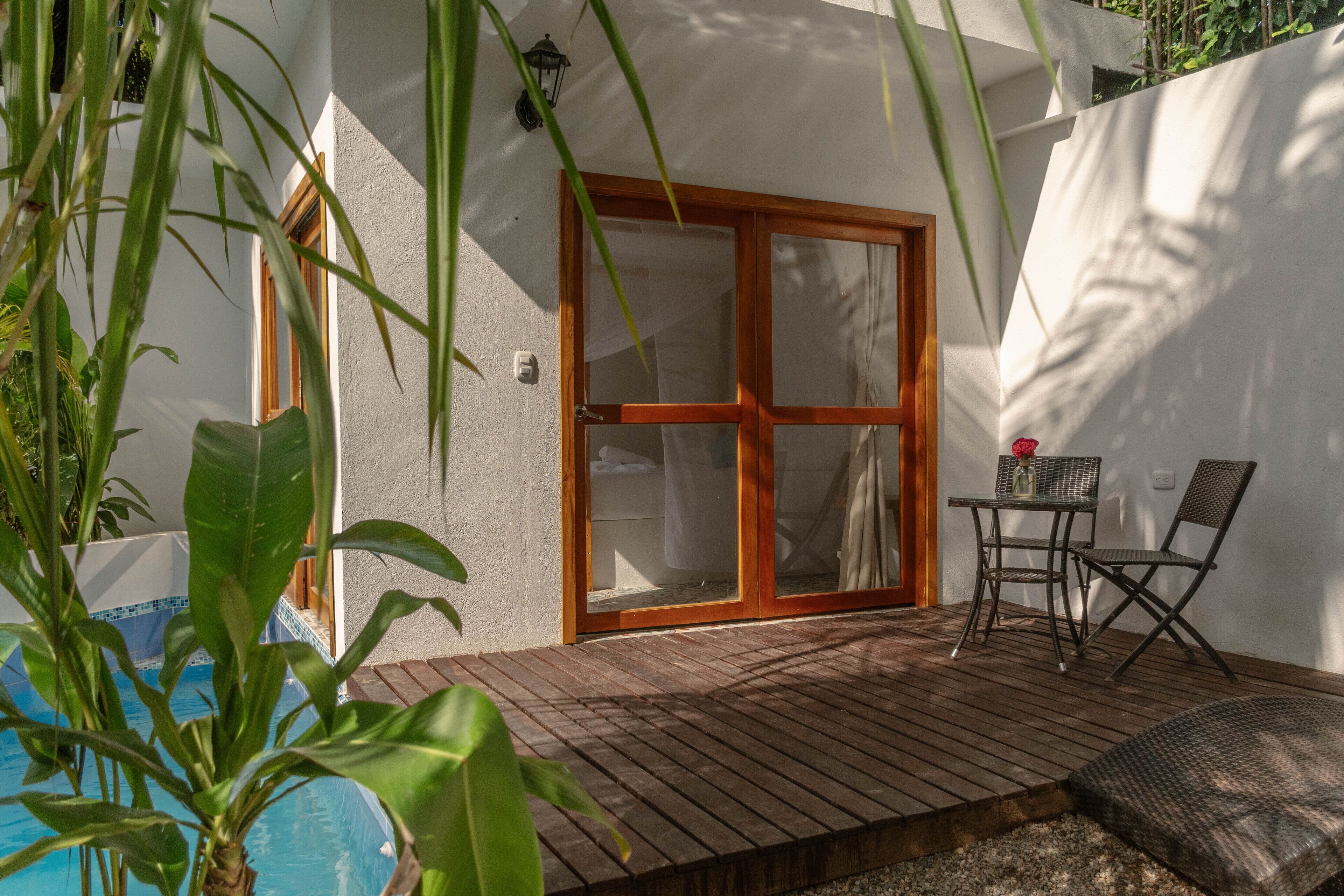 A wooden deck with a glass door leading to a pool area, surrounded by greenery.