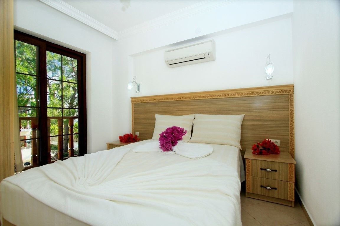 A neatly made bed with white linens and a decorative purple flower. There is a wooden headboard, a nightstand with a drawer, and an air conditioning unit on the wall.