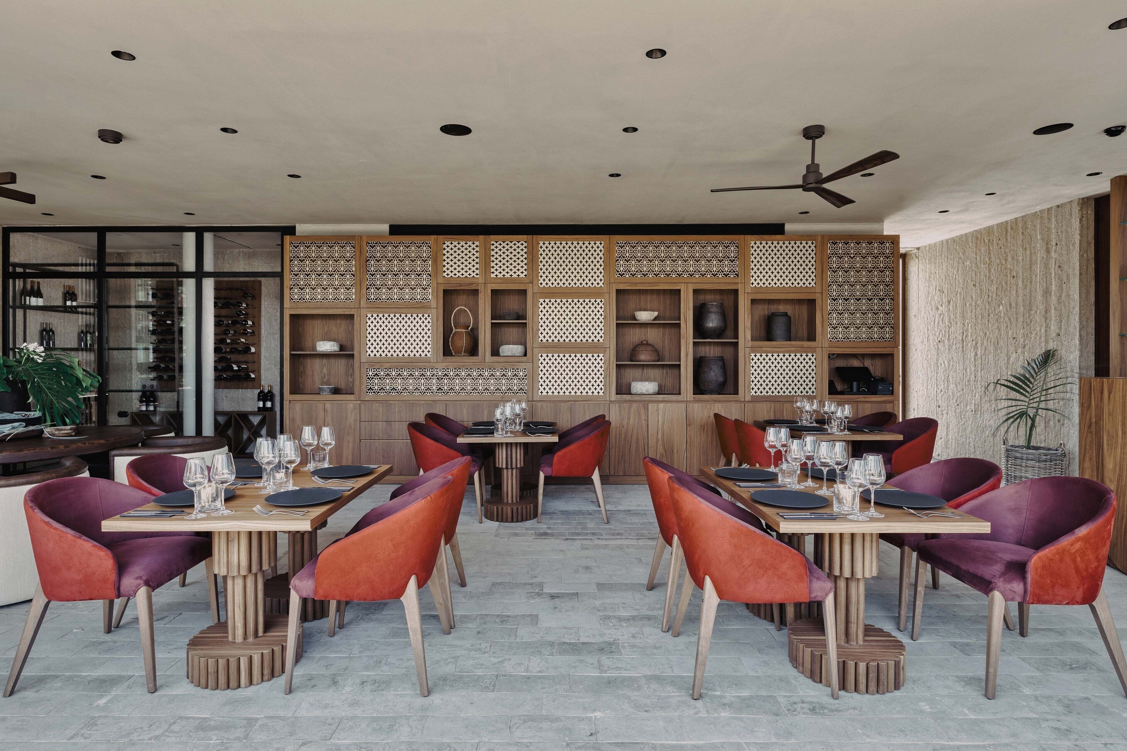 A restaurant with wooden tables and red chairs, a ceiling fan, and a decorative wall with shelves.