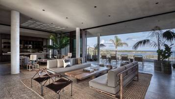 A spacious living area with a large sofa, coffee table, and chairs, a kitchen in the background, and a balcony with a view of the ocean.